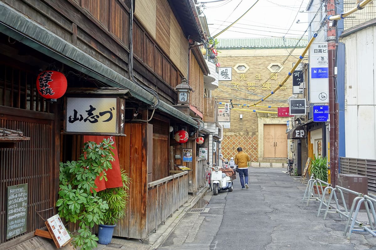 Looking down a narrow Pontocho alley towards the Pontocho Kaburenjo: the theatre's ochre facade carries its name in large Japanese characters above a diamond-patterned wall and closed wooden doors, while the alley is lined with wooden restaurant frontages, red lanterns and hanging signs; one person in a yellow jacket walks away from the camera.