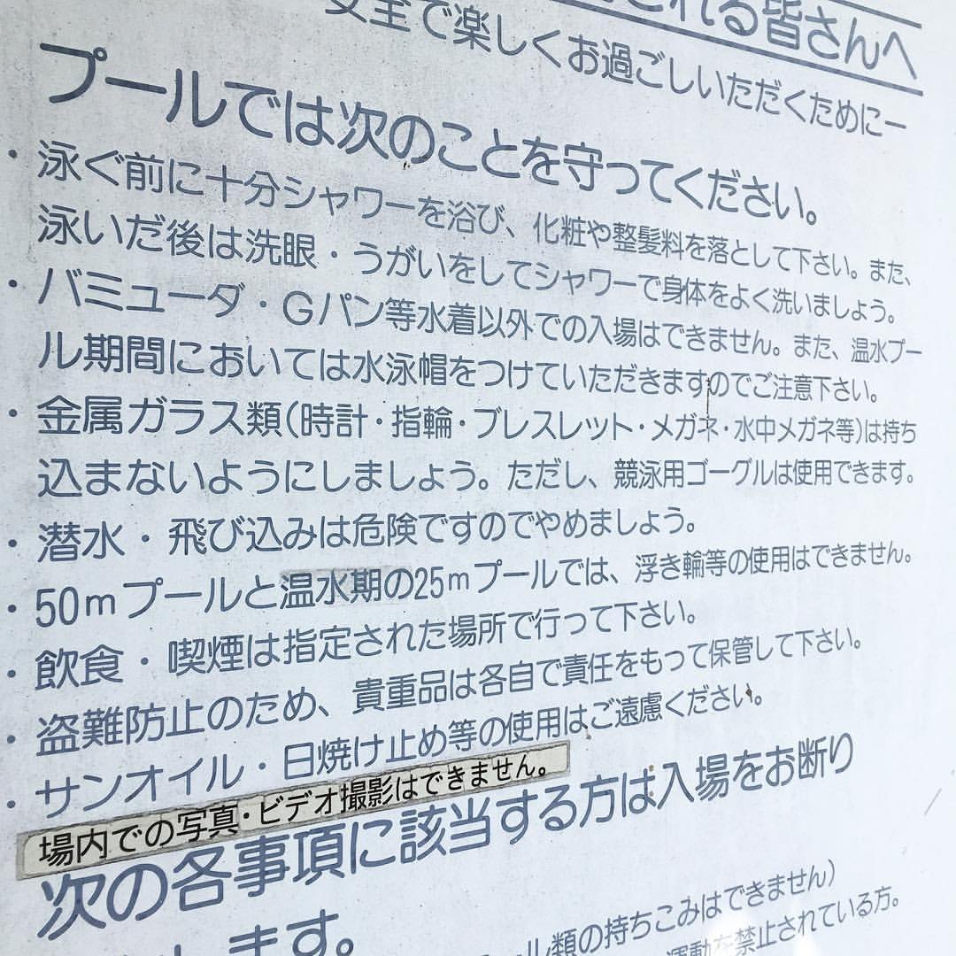 A close, angled shot of a Japanese pool noticeboard headed "please observe the following at the pool", followed by a long bulleted list: shower thoroughly before swimming, no Bermuda shorts or jeans in place of swimwear, swim caps required in the heated-pool season, no metal or glass items, no diving, no floats in the 50-metre pool, eating and smoking only in designated areas, no sun oil; a separate strip below reads that photography and video are not allowed on the premises, and the bottom line begins the list of people who will be refused entry