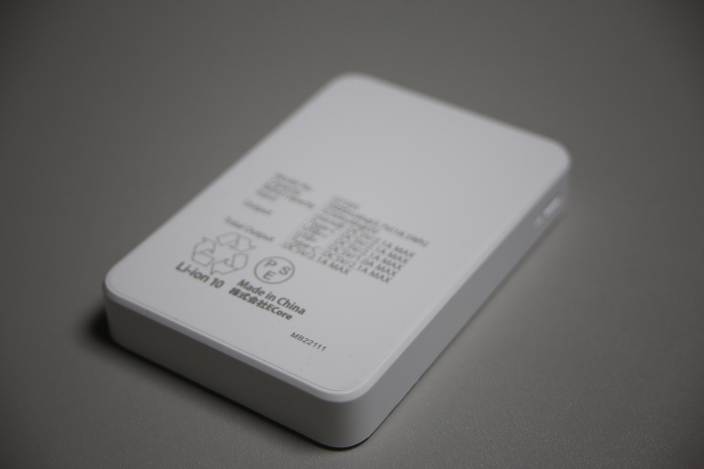 A white rectangular power bank lying face down on a grey desk, model number MB22111 printed at the lower right, a recycling triangle, a circular PSE mark and the words "Li-ion 10" and "Made in China" at the lower left, and a block of very fine specification text across the upper middle of the casing.