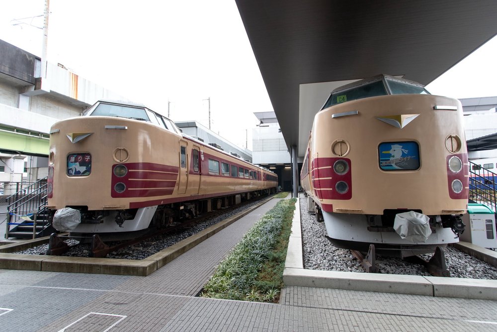 鐵道博物館戶外並排停著兩節奶油色車身、紅色腰帶的 183 系特急電車,左邊車頭的行先標寫「踊り子」,右邊車頭是海鷗圖案的「ライナー」標誌;兩車中間是一條種著矮灌木的步道,右邊那節停在一片大屋頂下面,後方是高架橋與館舍