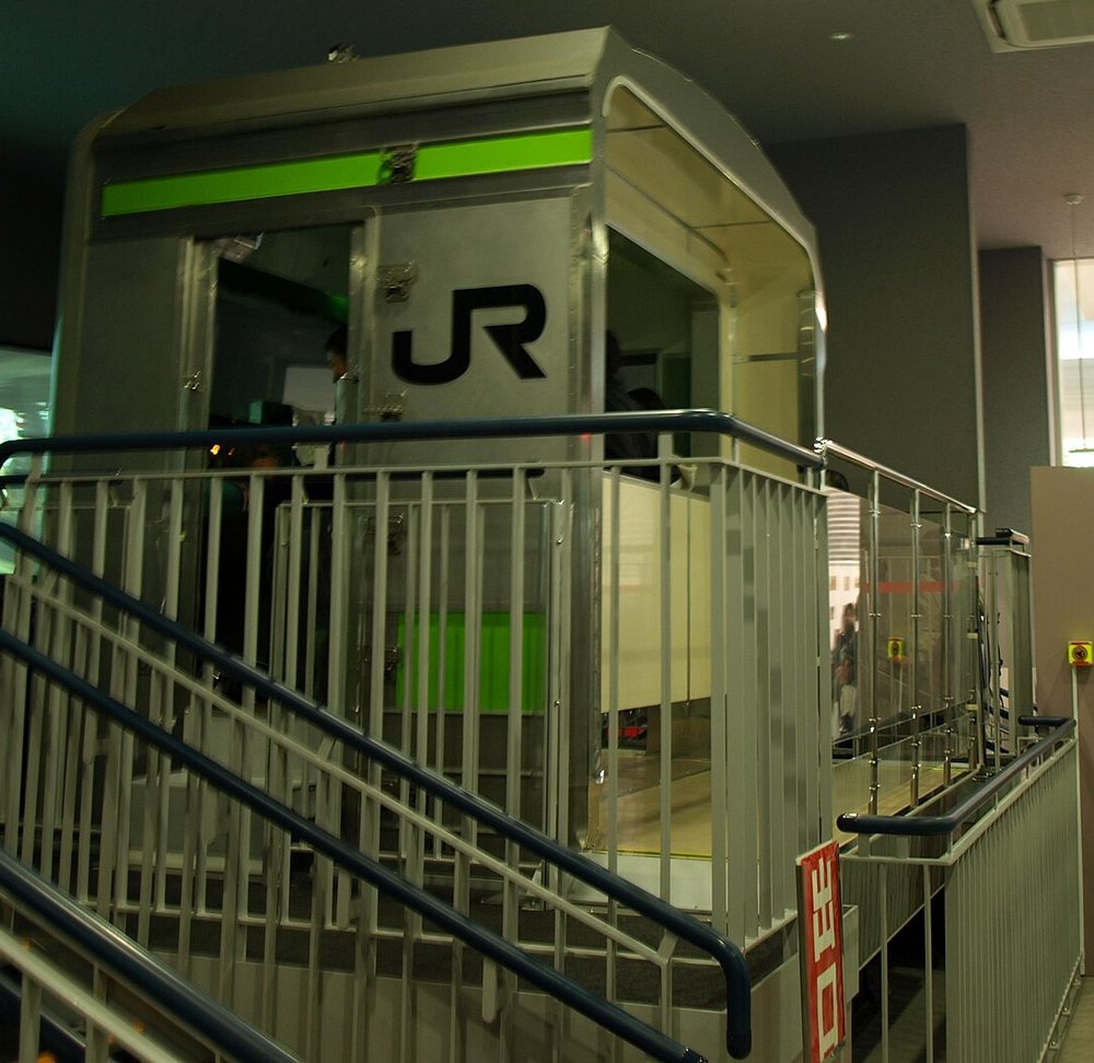 鐵道博物館的 205 系通勤電車模擬器:一個銀色車身、黃綠色腰帶、印著黑色 JR 標誌的電車駕駛室模型架在平台上,前面有藍色扶手的金屬樓梯通上去,駕駛室裡光線昏暗,右下角立著紅色直式的出口牌