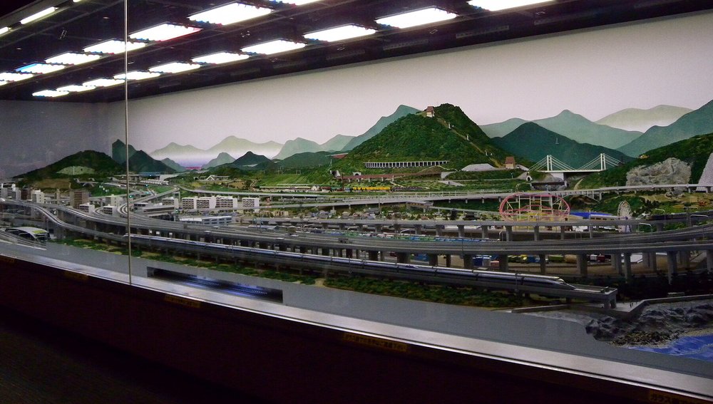 鐵道博物館的鐵道模型大廳:玻璃窗後面是一大片橫長的鐵道模型場景,前景是好幾層交錯的高架鐵軌,中間有住宅大樓、遊樂園的摩天輪,後方是綠色山丘、隧道、斜張橋與畫在牆上的遠山,天花板排滿日光燈