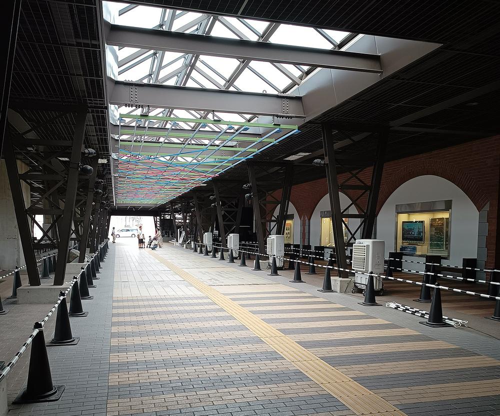 從鉄道博物館站往博物館入口的步道:頭頂是 New Shuttle 高架下的鋼骨與玻璃天窗,掛著一片綠、藍、紅色交錯的線條裝飾,右側是紅磚拱門牆面與展示櫃,地上鋪灰黃相間的磚與一條黃色導盲磚,兩側用黑色三角錐和鏈條圍出排隊動線