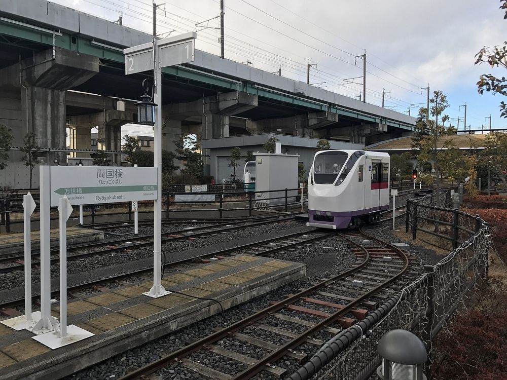 鐵道博物館戶外的迷你運轉列車軌道:一台白色車身、紫色裙邊的迷你特急列車停在彎道上,左邊是迷你月台與白底站名牌「両国橋 りょうごくばし」、往左寫「万世橋」往右寫「飯田町」,月台上立著號誌樁與一根吊燈柱;軌道兩旁是碎石與鐵柵欄,後方是新幹線的高架橋