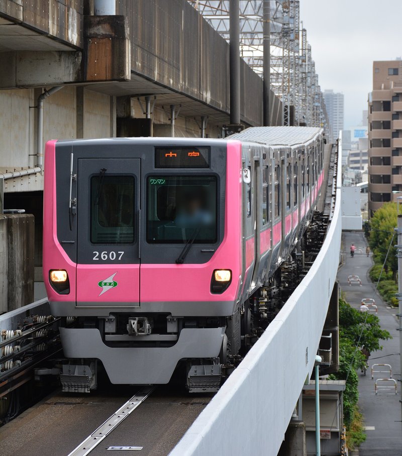 一列粉紅色車頭、銀色車身的 New Shuttle(埼玉新都市交通 2000 系)沿著高架軌道駛來,車號 2607,車頭上方的顯示器亮著橘色「大宮」,旁邊是 JR 新幹線的灰色高架橋,右下方是住宅區小路,駕駛員臉部已模糊處理