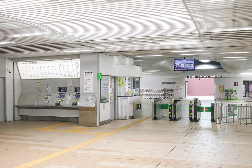 New Shuttle 鉄道博物館站的改札口:左邊牆上是「ニューシャトル旅客運賃表」與兩台綠色的「きっぷ・チャージ」售票機,中間是站務室窗口,右邊一排綠色箭頭的自動改札機,上方螢幕顯示往大宮與往內宿方向的下一班時間,遠處牆邊排著一整排扭蛋機