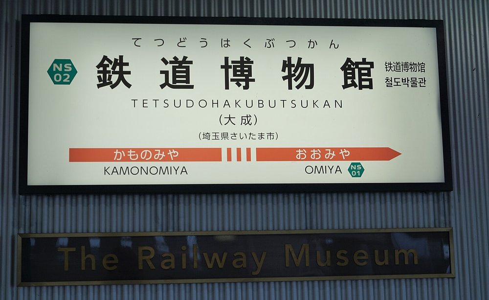 New Shuttle 鉄道博物館站的站名牌:白底黑字大大的「鉄道博物館」,上面有平假名讀音,左邊綠色六角形站號 NS02,下方寫 TETSUDOHAKUBUTSUKAN 與括號「大成」,最下面橘色箭頭往左是「かものみや」、往右是「おおみや NS01」;站名牌下方另掛一塊深色底金字「The Railway Museum」