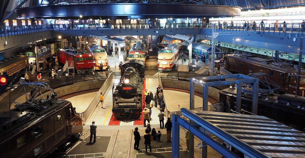 從二樓迴廊往下看本館的轉車台:圓形轉台中央停著黑色蒸汽機車 C57,車頭亮著燈、前面鋪紅地毯,轉台邊有十幾位參觀者站著看;後方一排是紅色電力機車、奶油色紅帶特急電車與藍色臥鋪客車,左下角是一台咖啡色電力機車的車頂與集電弓