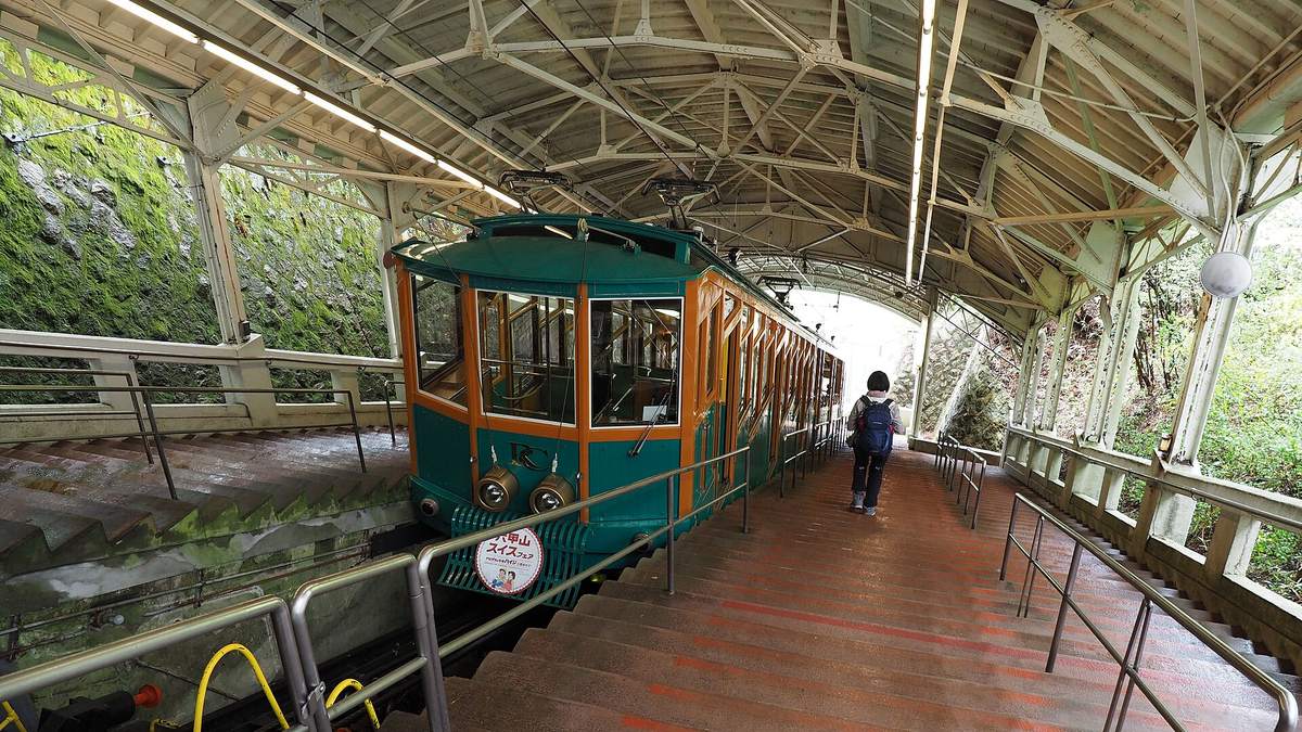 六甲纜車的車廂停在六甲山上站的月台：車身綠色配橘色窗框，車頭有兩盞圓燈與一塊圓形頭牌；月台是一整段往下的階梯，上方是白色鋼骨屋頂，一位背著背包的乘客背對鏡頭往下走