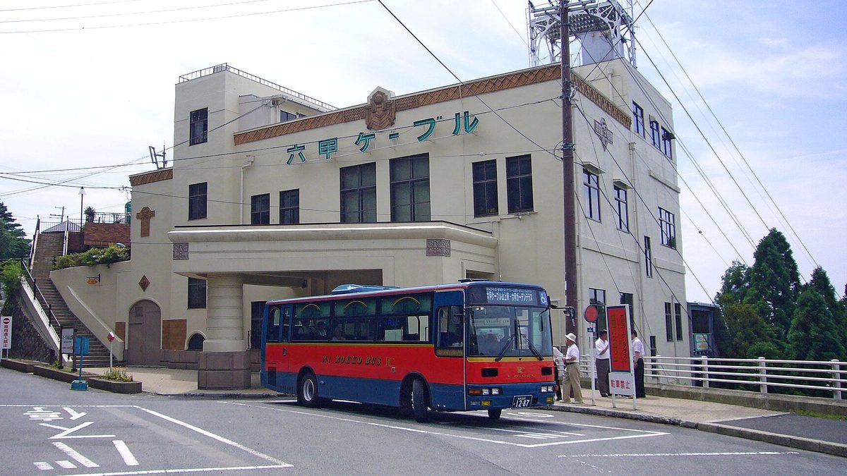 2006 年的六甲纜車山上站：米色三層樓站房外牆有綠色的「六甲ケーブル」字樣，站房正前方停著一台紅藍兩色的山上巴士，車頭方向幕寫「六甲ケーブル山上駅－六甲ガーデンテラス」，車旁有幾位乘客站在站牌邊