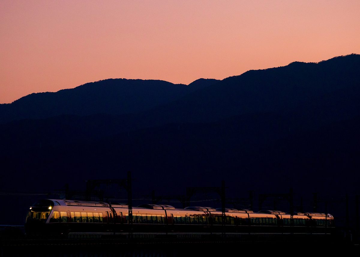日落後橘粉色天空下，一列 GSE 浪漫特快亮著車內燈駛過酒匂川鐵橋，背後是黑色的山稜線