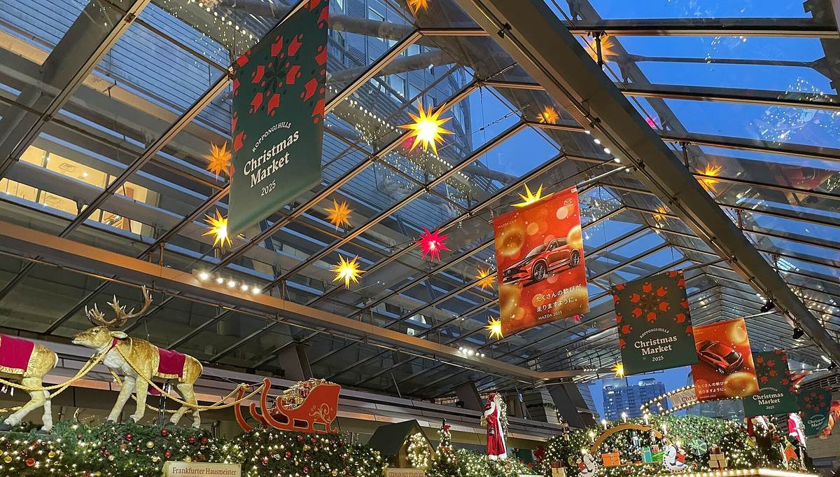 2025 年 12 月六本木之丘聖誕市集:玻璃大屋頂下掛著綠底的「ROPPONGI HILLS Christmas Market 2025」布旗與黃色、紅色的星形燈,下方木屋攤位的屋頂上鋪滿綠葉與燈串,站著馴鹿、雪橇與聖誕老人的裝飾