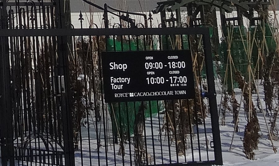 館外黑色鐵柵門上的營業時間牌：Shop 09:00–18:00、Factory Tour 10:00–17:00，下方小字最終入場 15:00，底部印著 ROYCE' CACAO & CHOCOLATE TOWN