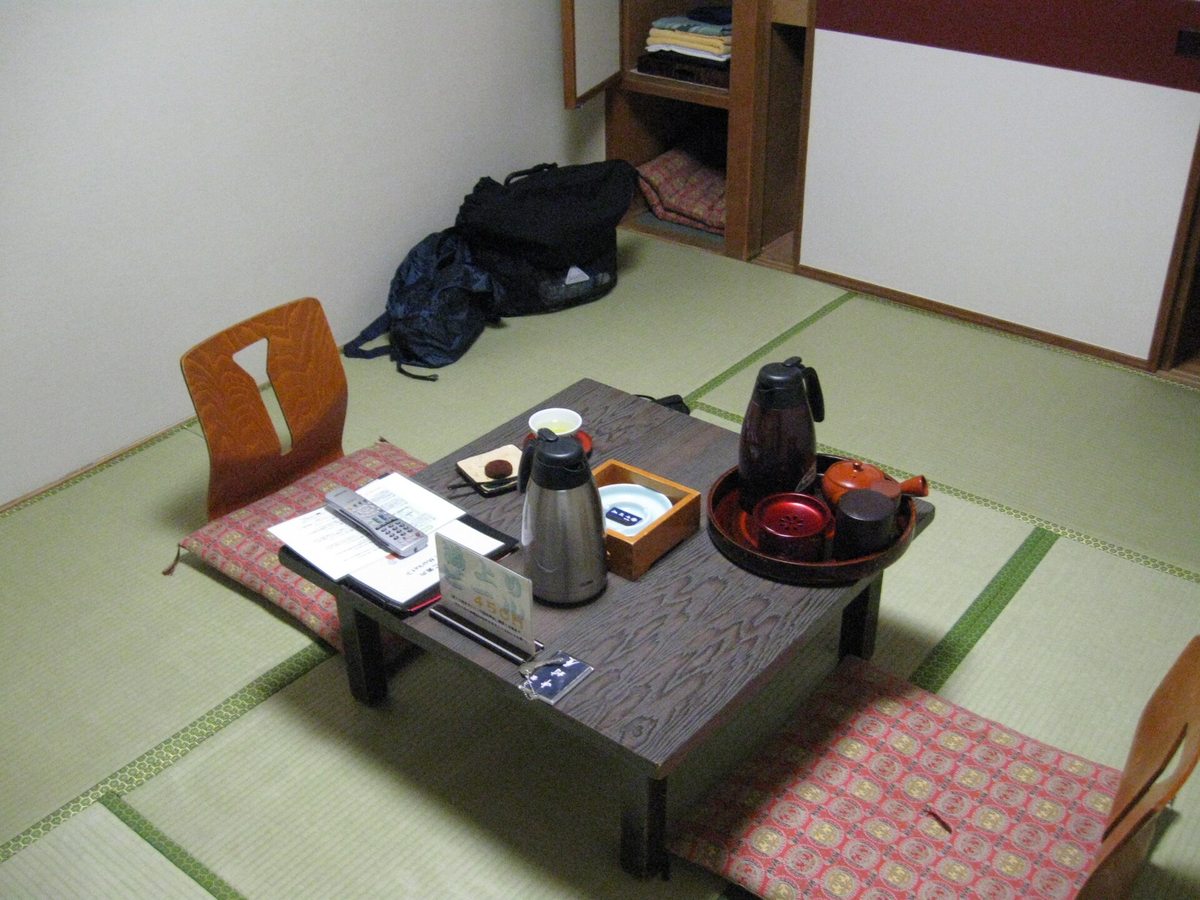 A tatami guest room in a Japanese inn: a low table in the centre set with a tea service, vacuum flask and remote control, floor cushions and legless chairs on either side, and an open closet behind stacked with yukata and spare cushions