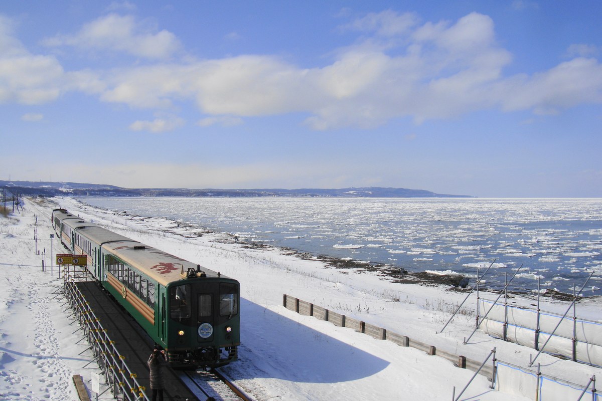 Drift ice on the Sea of Okhotsk: pale floes packed across grey-blue water right up to the shoreline, with a snow-covered railway embankment along the coast