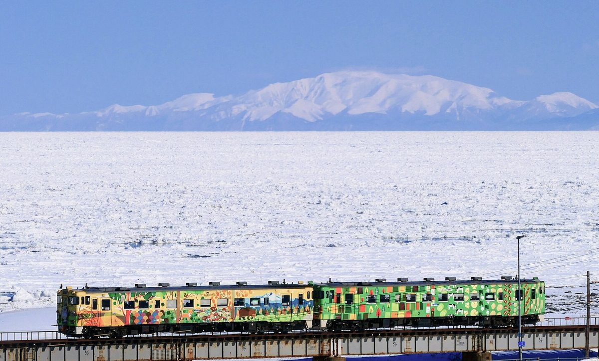 The Ryuhyo Monogatari: two painted diesel railcars, one yellow-orange and one green, crossing a girder bridge with the Sea of Okhotsk completely covered in drift ice behind and the snowy mountains of the Shiretoko Peninsula in the distance
