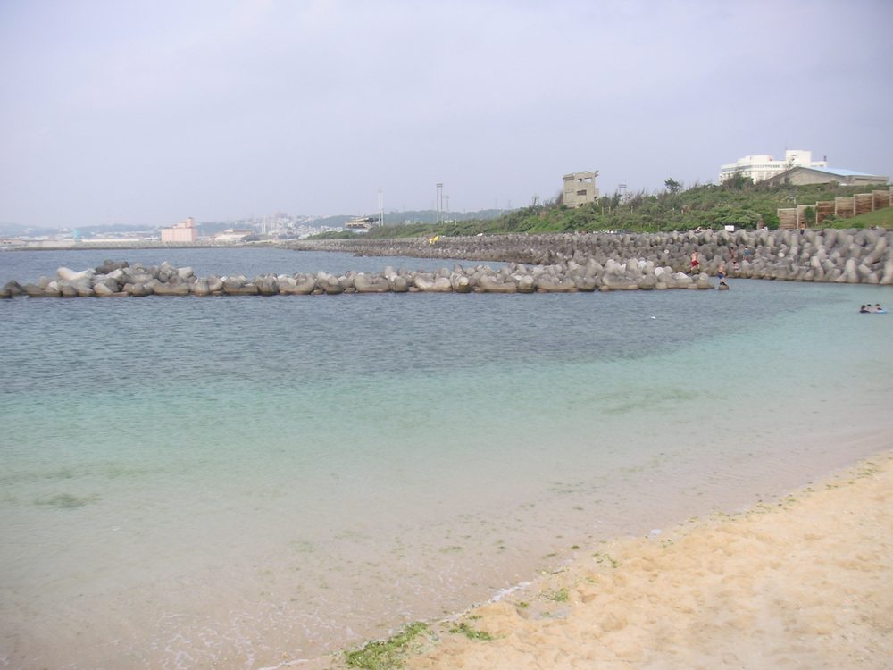 陰天白天的宜野灣トロピカルビーチ,前景是淺綠色的淺水與沙灘,海面被一排灰色消波塊防波堤圍起來,遠處是宜野灣的海岸線與建築
