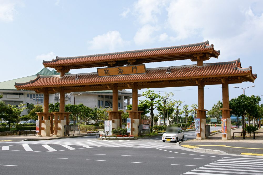 宜野灣海濱公園入口的歓海門,兩層紅瓦屋頂、木柱撐起的大型門樓,中央匾額寫著「歓海門」,門前是斑馬線與一輛白色小車,後方是綠屋頂的建築與行道樹