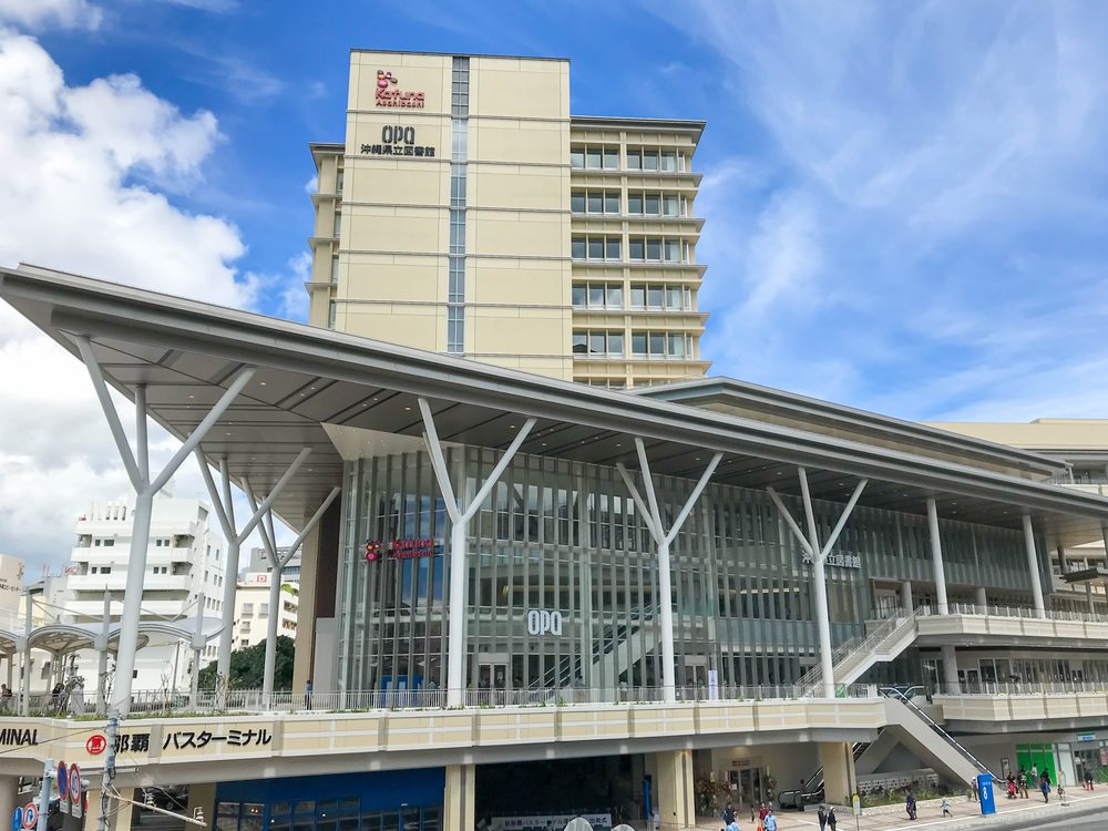 晴天的那霸巴士總站外觀,白色 Y 字形柱子撐起大片屋簷,下方招牌寫著「那覇バスターミナル」,後方米色大樓上有 OPA 與沖縄県立図書館 的字樣