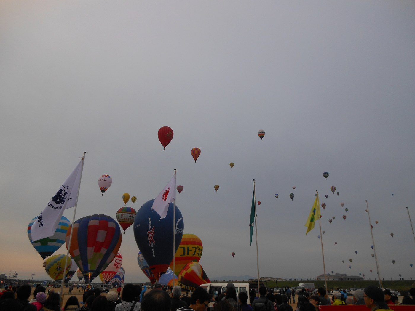 Dawn on the riverbank: the bottom of the frame is a solid mass of spectators seen from behind, while dozens of hot air balloons climb away and scatter across the grey sky between flagpoles