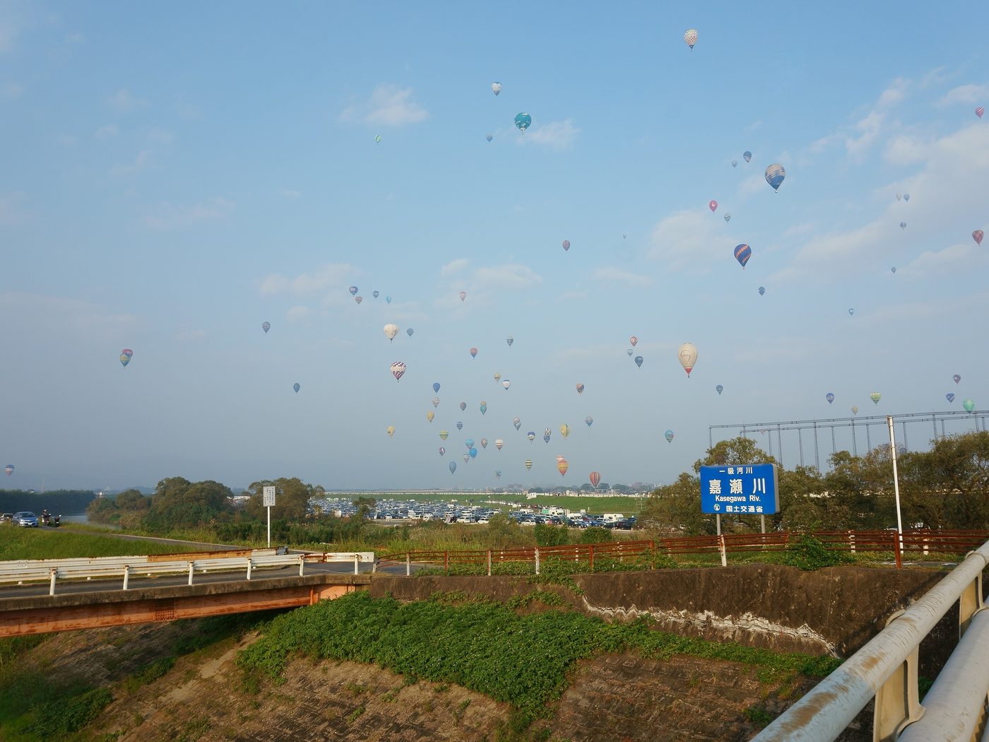 Seen from a bridge over the Kase River: dozens of hot air balloons scattered across a blue sky, an unbroken line of parked cars below the levee, and a blue roadside sign reading Kasegawa Riv.