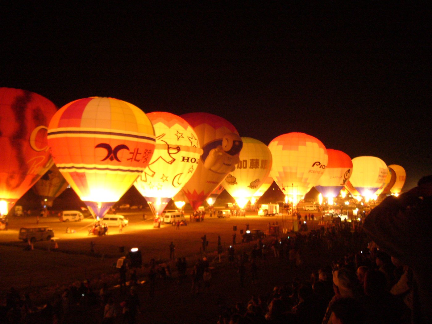 A row of tethered hot air balloons at night, envelopes glowing orange from the burners inside, support vehicles parked along the ground and a dense crowd silhouetted in the foreground