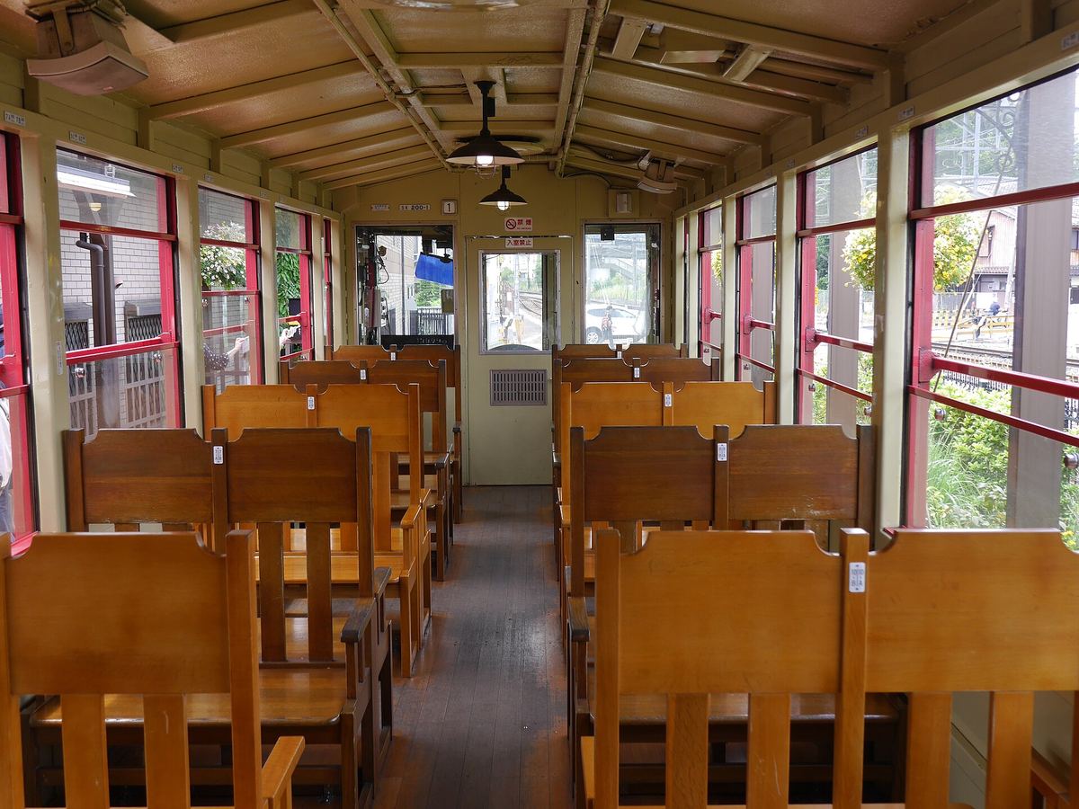 嵯峨野小火車現行車 1 號車車內：沒有軟墊的木椅四人對坐排在走道兩側，兩側是紅框的車窗，天花板吊著燈，車廂盡頭是駕駛室的門