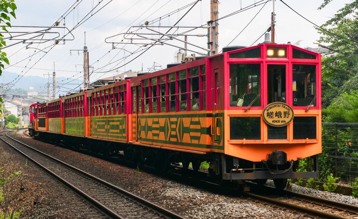 2026 年 7 月的嵯峨野小火車現行車：最前面是紅橘兩色、掛著「嵯峨野」圓牌、有駕駛室的 1 號車，後面接著紅色與黃色的客車，隊伍最尾端是紅色的機關車，行駛在架線下的彎道上