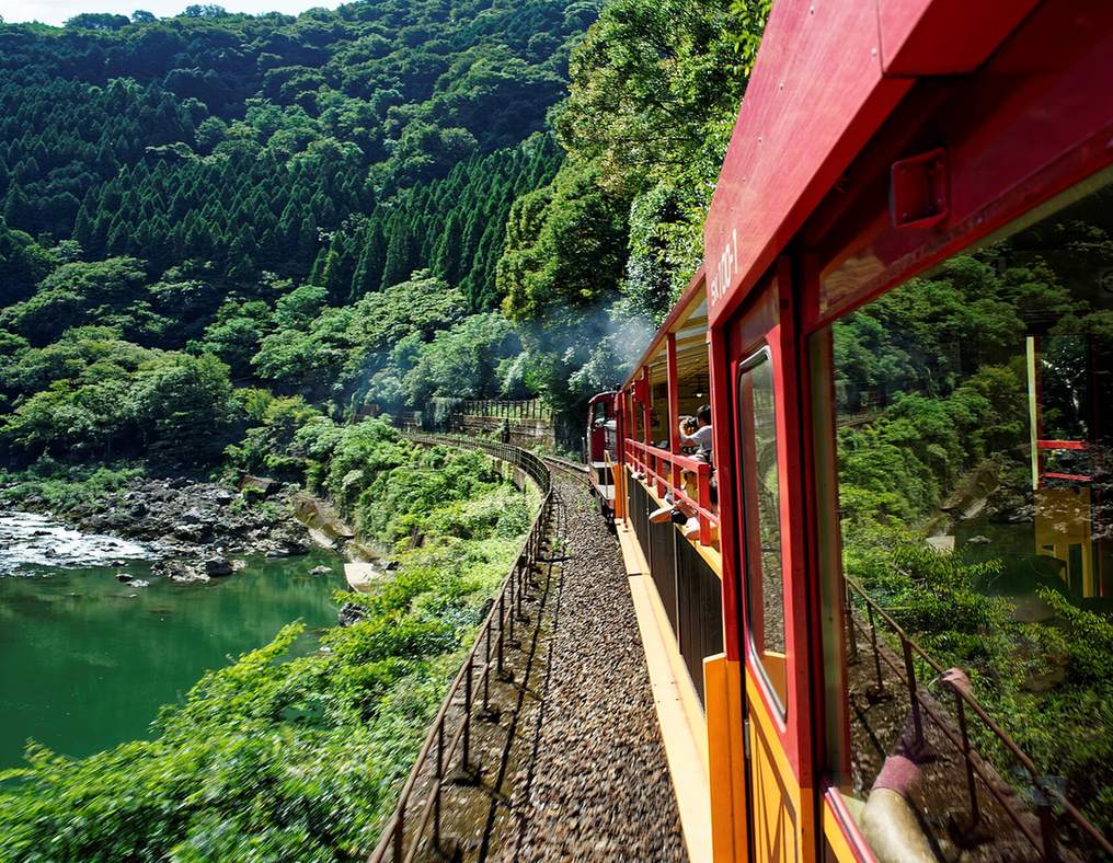 2016 年 8 月從嵯峨野小火車車上往前看：紅色車廂沿著彎道前進，前面的車廂有乘客把相機伸出欄杆外，左下方是墨綠色的保津川與岩石河岸，最前頭的機關車冒著白煙