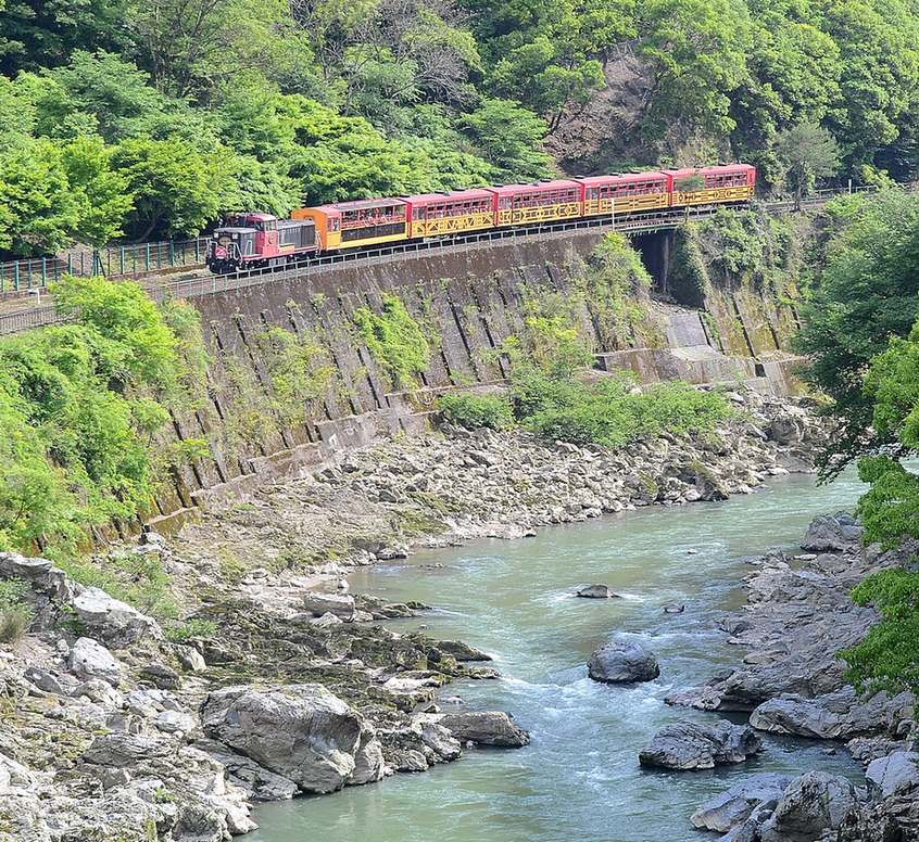 2016 年 5 月新綠的保津川溪谷：機關車與五節紅黃色客車的嵯峨野小火車沿著河岸山壁上的軌道行駛，下方是岩石河床與青綠色的保津川