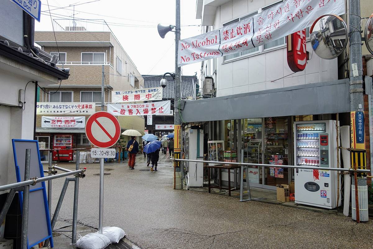 雨天的商店街街口，路面濕亮：橫掛在半空的白色布條印著「No Alcohol／No Tattoos」與中文「禁止饮酒／禁止纹身」及韓文；前方另一條紅字布條寫「酒気・いれずみ・地下足袋・履物禁止 検問中」，其下白底黑字寫「午後7時30分以降 一般客入場禁止」；左側看板寫「北門 西門から入場してください」，路中央立著一面禁止進入的圓形交通標誌，下方時間牌寫 15:20-19:30；街道深處有幾名撐傘的行人背影走遠