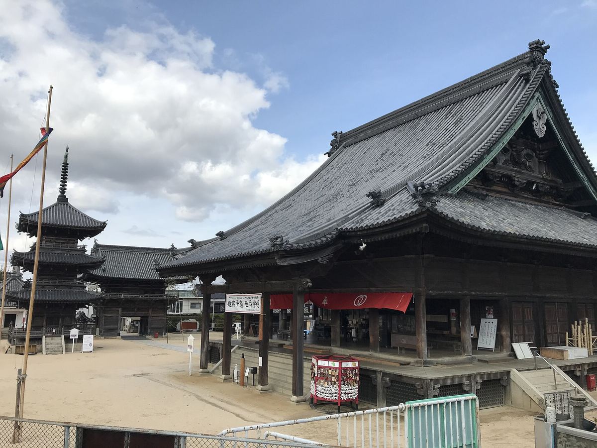 西大寺観音院的本堂近景：巨大的灰瓦屋頂與大幅翹起的簷角，正面木柱之間垂著一整條紅色布幕，柱前是抬高的木造平台與一具紅色的賽錢箱，庭中鋪著沙土，左後方是三重塔與寺務所，天空有積雲