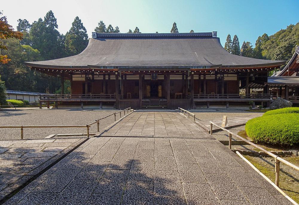 西芳寺本堂(西來堂)的正面:大片灰色屋頂的木造佛堂,前方是一條石板參道與竹製欄杆,右側是修剪成圓球的灌木,晴天