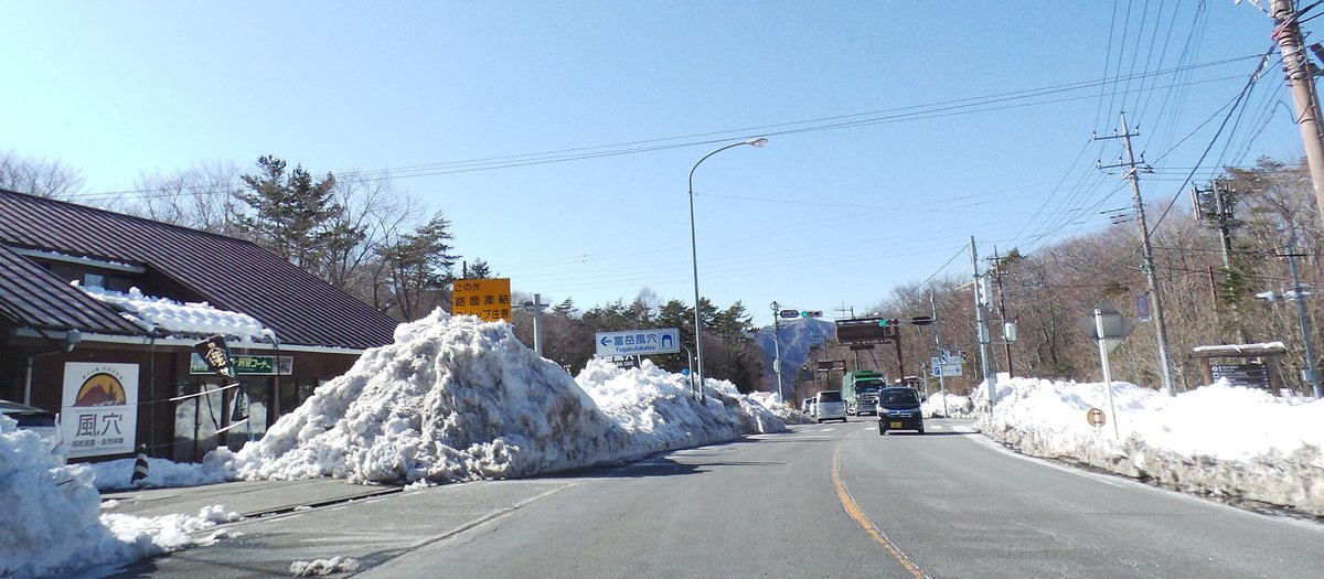 西湖南側往富岳風穴的路口附近:兩側路邊堆著比車還高的除雪雪堆,黃色告示牌寫「この先 路面凍結 スリップ注意」,藍色指標寫「富岳風穴」往左