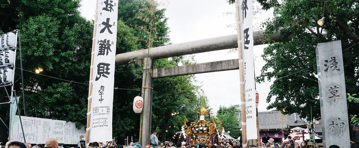 淺草神社的石造鳥居，兩側立著白底黑字的長幡，右邊石柱刻著「淺草神社」，鳥居後方一座金色神輿露出屋頂，下緣是人群的頭頂