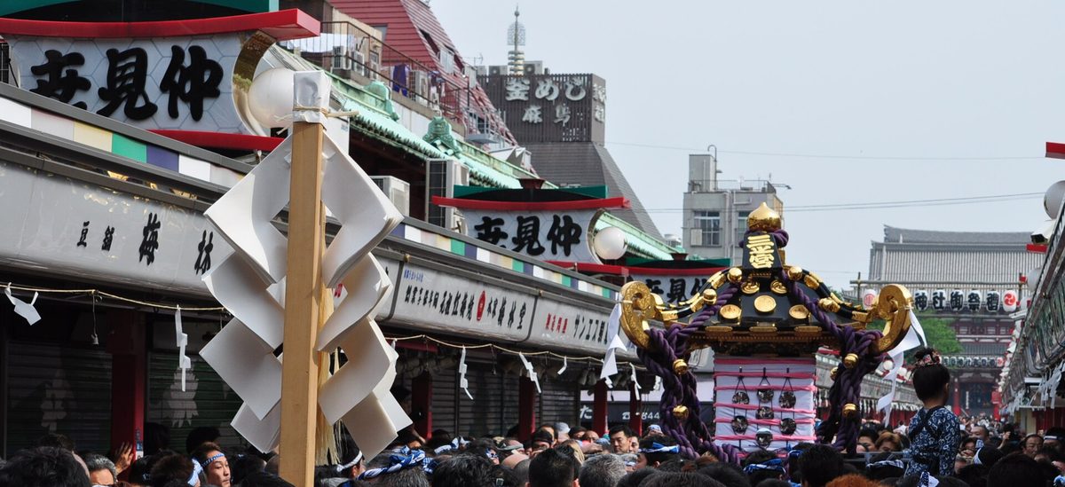 仲見世商店街裡的本社神輿：屋頂是黑漆金飾、頂端一顆金色寶珠，繫著紫色粗繩，周圍擠滿人；兩側店家的鐵捲門還拉著，招牌寫著「仲見世」