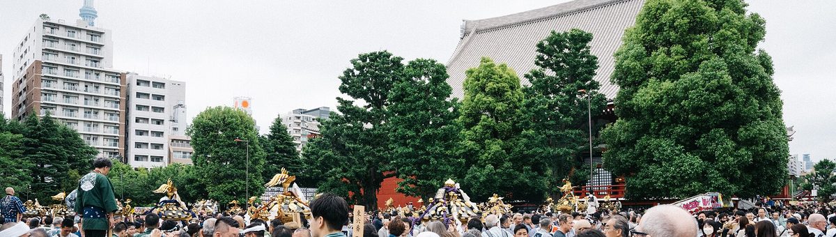 淺草寺本堂後方的廣場：樹叢後面露出本堂的大屋頂，樹下一字排開好幾座金色神輿的屋頂，前方是滿滿的人頭，左後方遠處看得到晴空塔