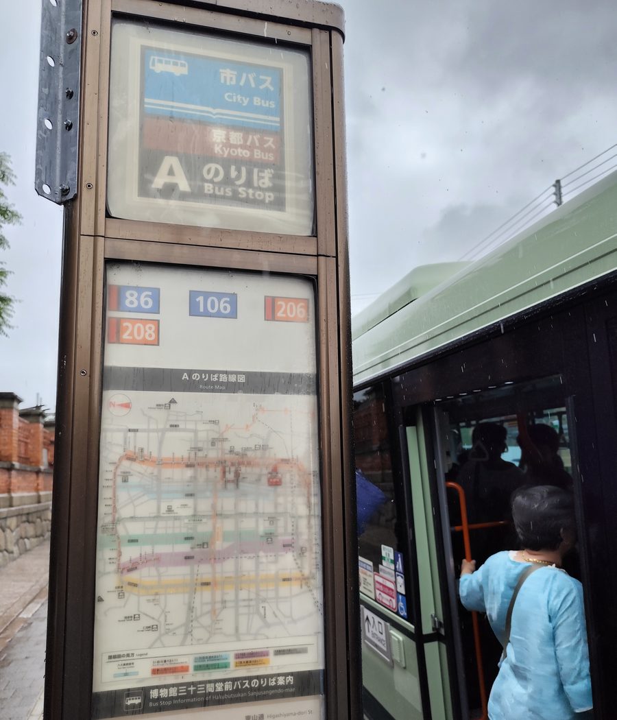 雨天裡「博物館三十三間堂前」A のりば的咖啡色站牌：上方寫著市バス City Bus、京都バス、A のりば，中段是四個系統號碼 86、106、206、208，下方是 A のりば路線圖，最底下一行「博物館三十三間堂前バスのりば案内」；右邊一輛淺綠色市巴士停靠，一位穿淺藍上衣的乘客背對鏡頭正要上車