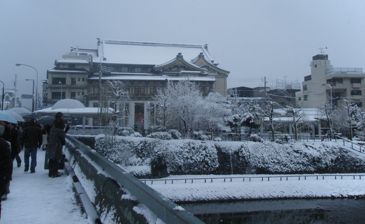 大雪中的京都四条大橋東端：左邊橋上的人行道積著雪，幾位穿深色外套、撐傘的行人背對鏡頭往前走；正前方是屋頂鋪滿白雪的南座，鴨川河岸的樹木與矮樹叢都被雪蓋住，天空灰白一片