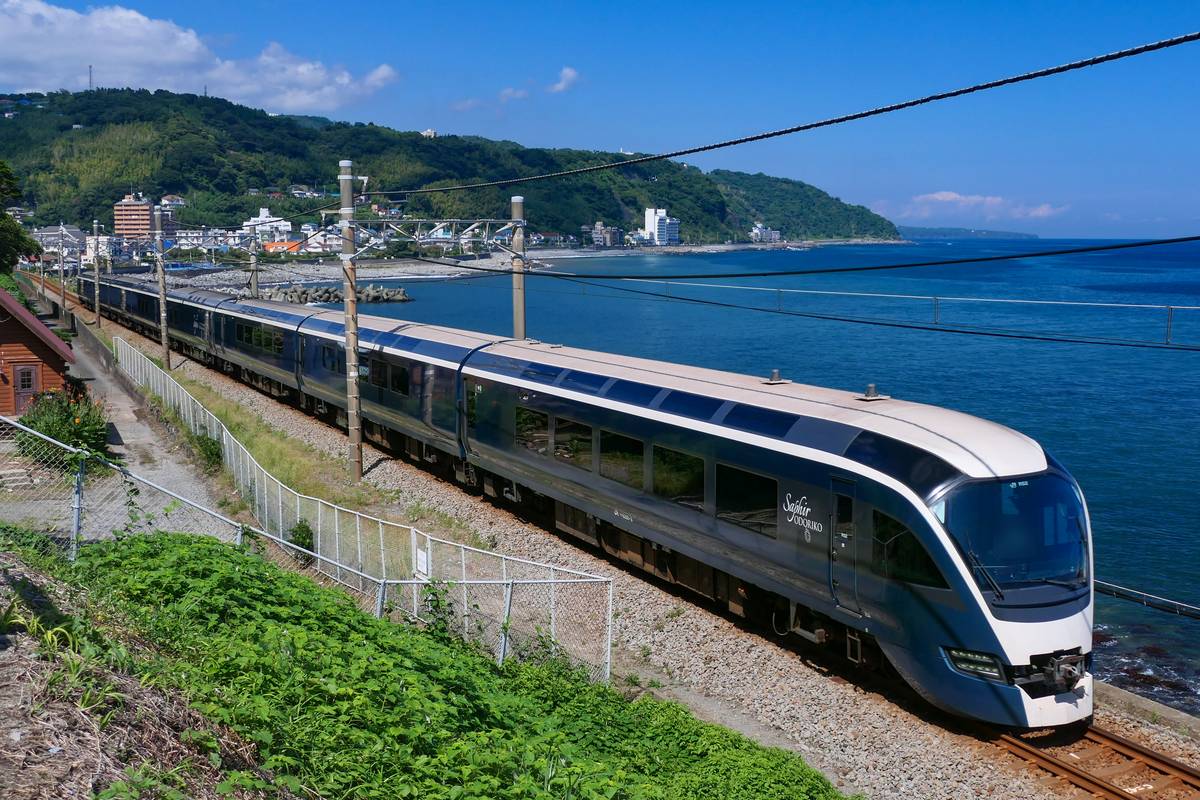 SAPHIR 踴子號沿著伊豆半島東岸的海邊行駛:深藍色車身與連到車頂的大窗,右邊緊貼著深藍色的海,遠處是山坡下的海岸小鎮與白色大樓
