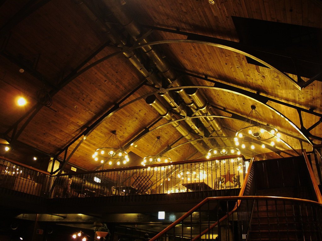 札幌啤酒園餐廳內部往上看:深色木板拼成的挑高斜屋頂,幾道彎曲的鐵拱架橫過天花板,下面吊著一圈圈小燈泡的吊燈,二樓是一整圈木欄杆圍起的迴廊座位