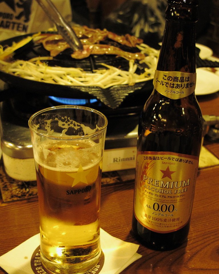 札幌啤酒園桌上的成吉思汗鐵鍋近景:中間隆起的黑色鐵鍋下面是藍色瓦斯火,鍋上烤著羊肉,周圍鋪滿豆芽菜,有人用夾子在翻;前面是一杯印著金色星星與 SAPPORO 的啤酒杯和一瓶 Sapporo Premium Alcohol Free 無酒精啤酒,左上角看得到「BIER GARTEN」字樣
