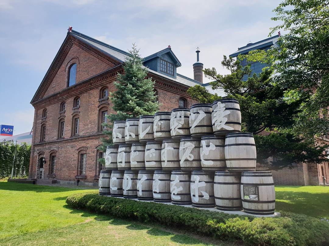 夏天的札幌啤酒博物館側面:紅磚建築前的草地上,木酒桶疊成金字塔形的牆,每個桶身寫著一個白色大字,由右往左直讀成一句古日文標語;旁邊有一棵高大的藍杉