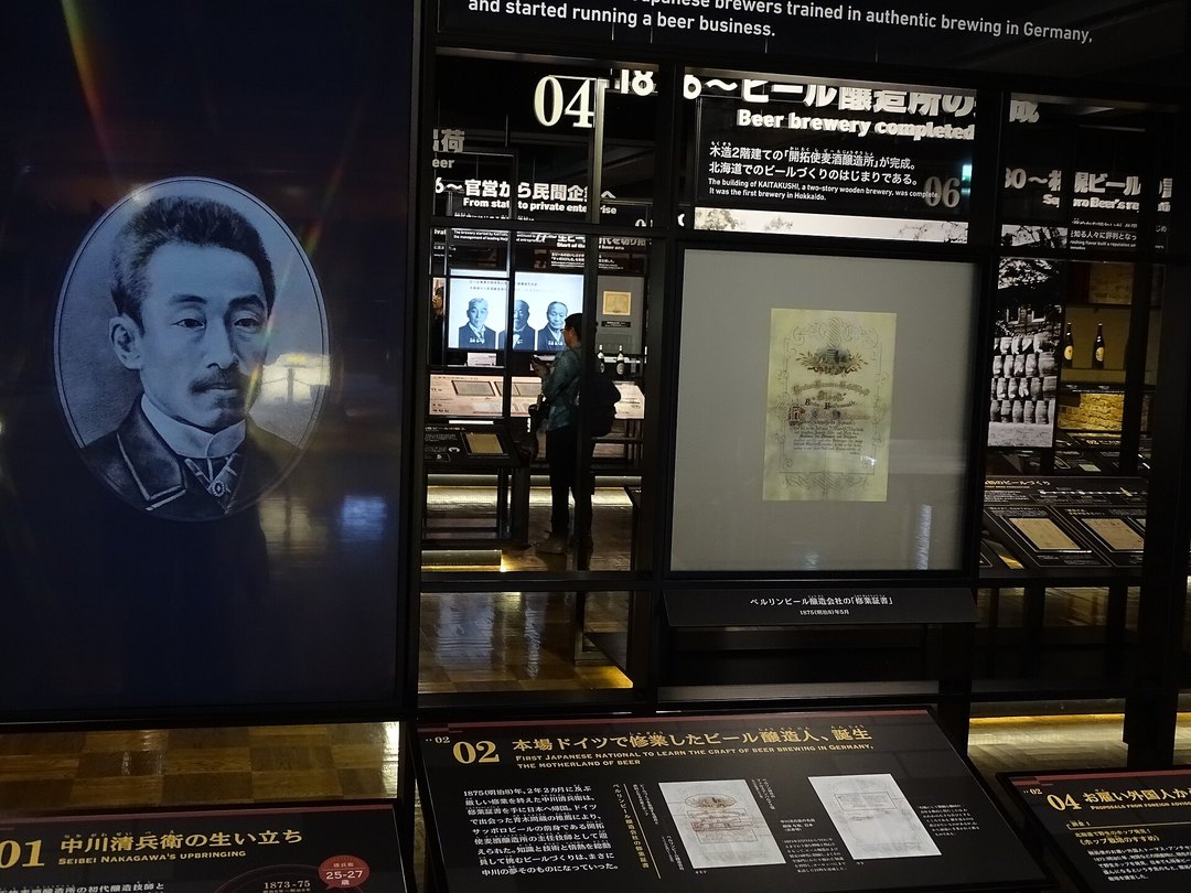 札幌啤酒博物館的展示區:左邊是一位留八字鬍男士的黑白肖像,右邊黑色展板上編號 02、04 的日英雙語說明,寫著在德國學釀酒與「ビール醸造所」完成,中間玻璃框裡是一張舊文件,後方深處有一位參觀者背對鏡頭