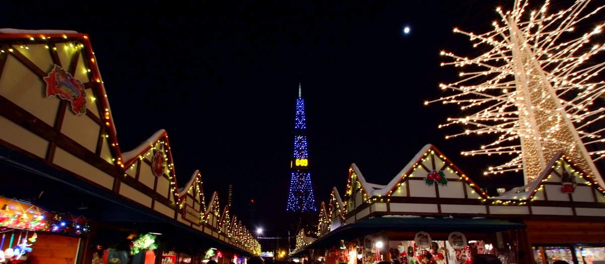 The Munich Christmas Market in Sapporo at night in December 2013: two rows of wooden chalet stalls with string lights along the eaves and snow on the roofs form an aisle, the Sapporo TV Tower lit blue straight ahead and a white tree-shaped light sculpture at upper right