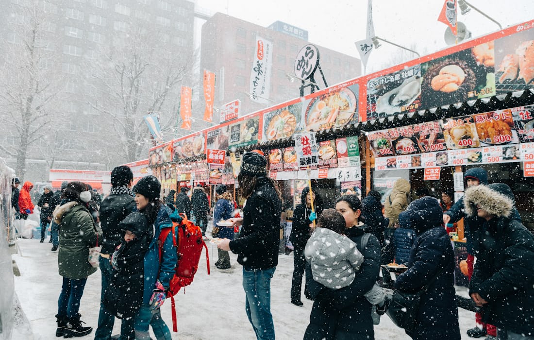 A downtown Sapporo food-stall street during snowfall with bundled-up crowds