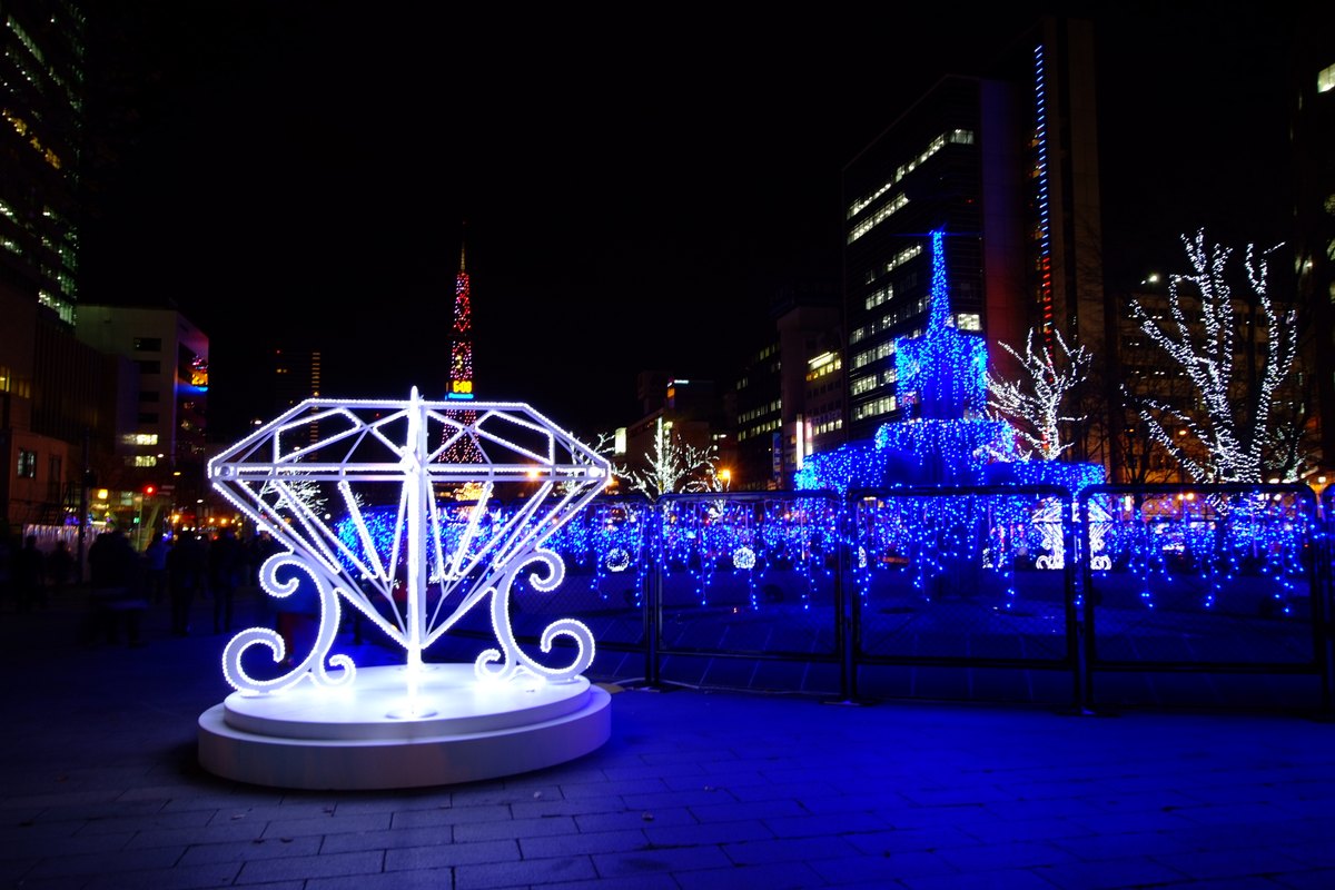 Sapporo White Illumination at Odori Park in November 2015: a white LED diamond-shaped sculpture in the foreground, a fountain wrapped in blue string lights on the right, white-lit trees behind and the Sapporo TV Tower lit red in the distance