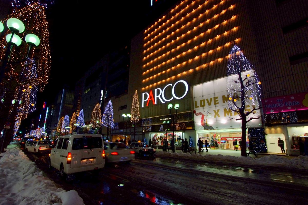 Night street scene in front of Sapporo PARCO in December 2013: a row of roadside trees covered in gold and blue-white string lights, the PARCO facade lit with rows of orange lamps, snow along the road and taxis queuing