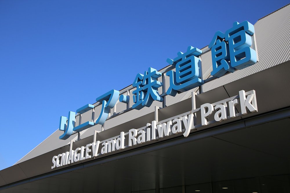 從下往上拍的入口雨遮,上面立著藍色立體字「リニア・鉄道館」,下面一排白色立體字「SCMAGLEV and Railway Park」,背景是藍天和灰色波浪板屋頂