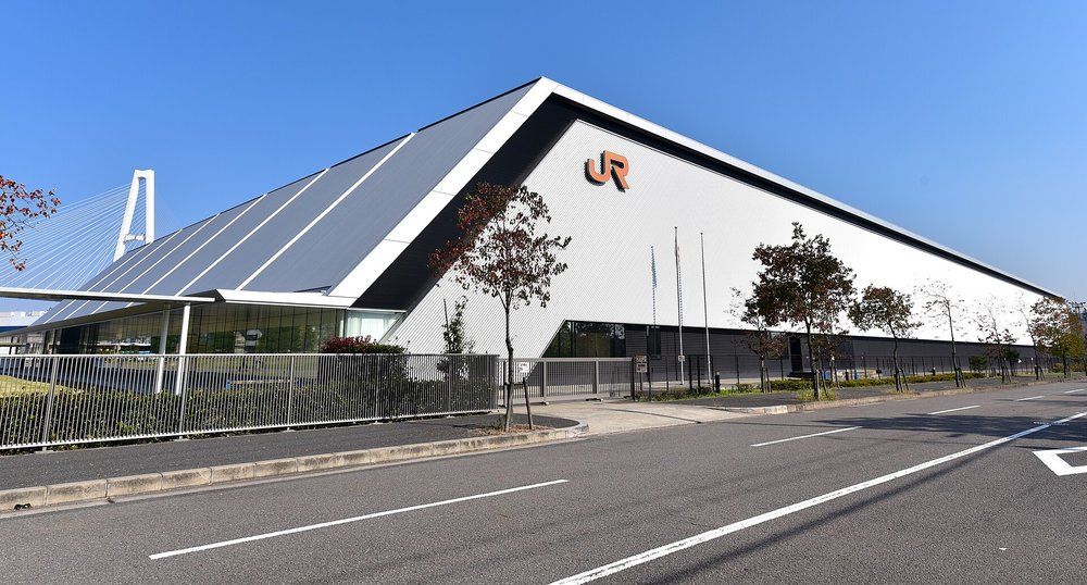 藍天下的磁浮鐵道館外觀:一棟白色與灰色的大型斜屋頂建築,三角形的白牆上掛著橘色 JR 標誌,前面是一排低矮行道樹、鐵欄杆和空曠的雙向車道,左後方露出名港中央大橋白色的斜張橋塔